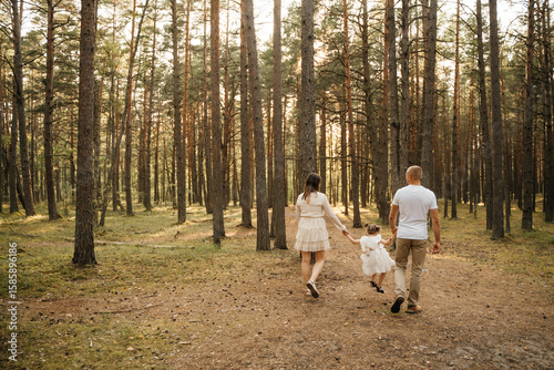 
A happy family of mother, father, and daughter walks hand-in-hand away from the camera on a sun-dappled forest path.