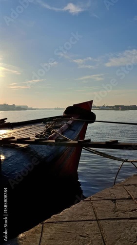 The bow of a wooden boat tied to a dock, with the setting sun creating beautiful reflections on the calm water