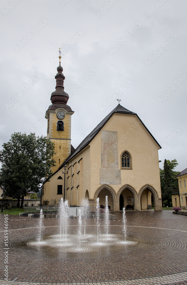 Naklejka premium water fountain in front of church in historic downtown of tarvisio italy europe (european travel destination near udine) rainy wet stormy rain weather history tourism