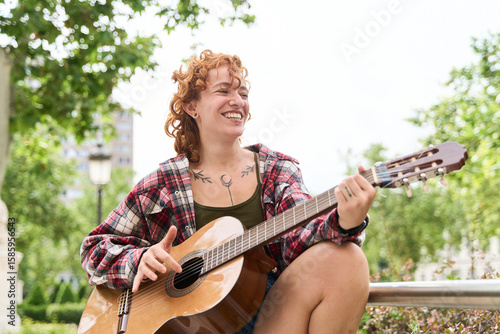 Canvas Print Young red haired musician happily playing acoustic guitar in a city park