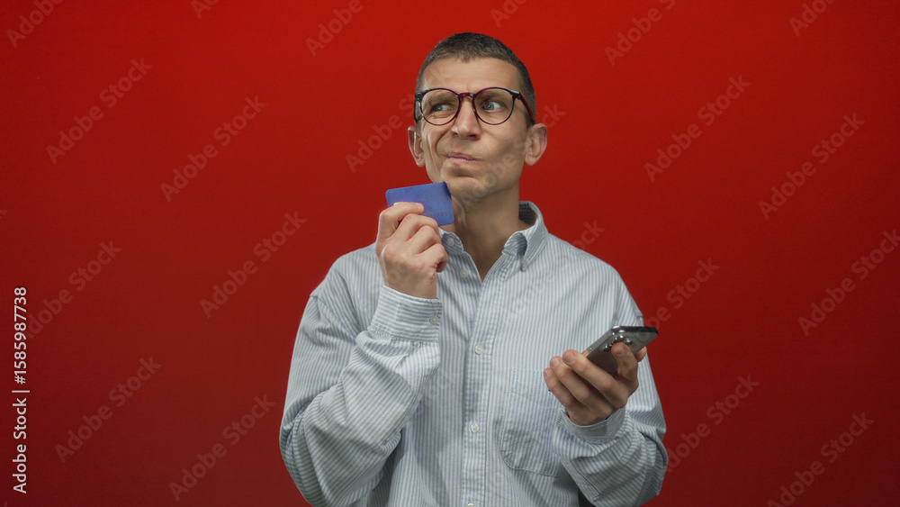 Fototapeta premium Man holding credit card and smartphone against a vivid red background, suggesting a financial decision or online shopping scenario with focus on technology and finance.