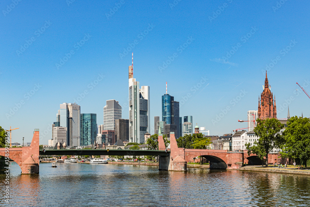 Fototapeta premium A Frankfurt city skyline in Germany with a bridge over a Main river. The bridge is red and the water is calm. Against the background of the blue sky.