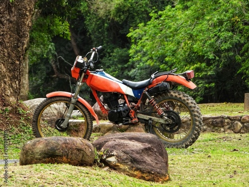 Orange and blue dirt bike parked on grassy terrain with large rocks and lush green trees