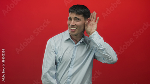 Tableau sur toile Man holds hand to ear listening against red backdrop in studio with inquisitive expression; curiosity