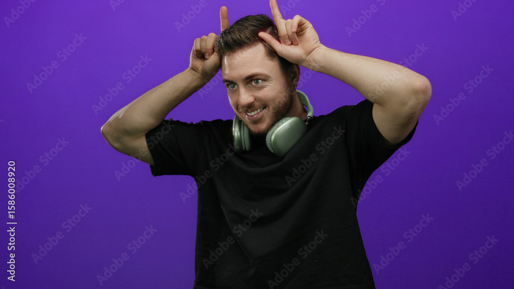 Naklejka premium Young man with beard wearing headphones makes playful horn gesture against purple wall in isolated setting, showcasing fun, isolated, casual, expressive, playful, and modern vibe.