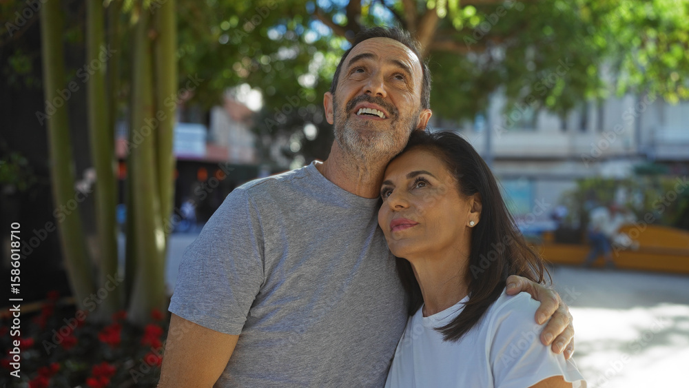 Fototapeta premium Middle-aged hispanic couple embraces lovingly outdoors in sunlit urban park setting, capturing romance and family connection between man and woman against city backdrop.