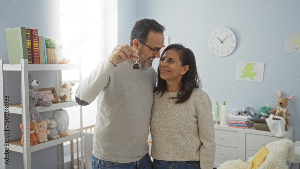 Fototapeta premium Middle-aged hispanic couple in bedroom holding keys, symbolizing new home and family with nursery decor in background, both smiling and embracing in warm indoor setting.