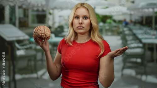 Cuadro en lienzo Woman in restaurant holding brain wearing red shirt with confused expression, surrounded by tables under white umbrellas