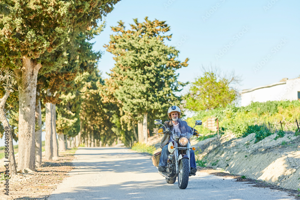 Obraz premium Senior man riding motorcycle on countryside road with trees