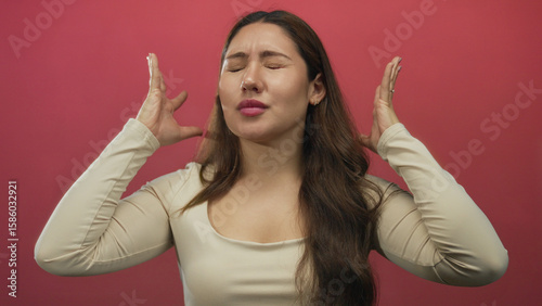 Woman holds head with hands in a pink studio setting showing closed eyes and tense expression; stress.