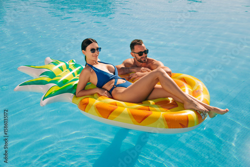 Fototapete Woman lying on a pineapple float in the pool, young handsome man swimming nearby