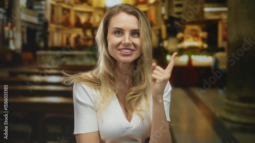 Young woman smiling inside a church with wooden benches and a bright background, showcasing her blonde hair and engaging expression in a peaceful religious setting.