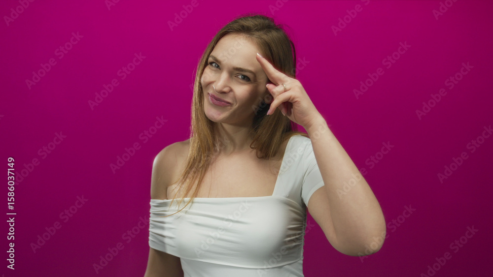 Young woman smiling with bare shoulder and pointing finger to temple in a studio with pink background; confidence. Young woman smiling with bare shoulder and pointing finger to temple in a studio with pink background; confidence.