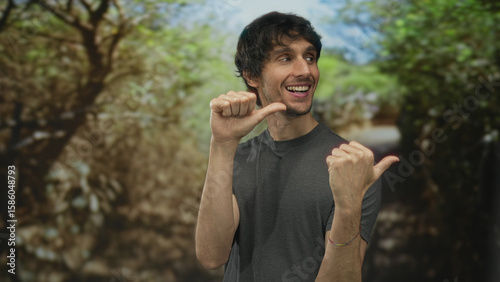 Fotografia Man in gray shirt thumbs pointing to chest on a sunlit green forest path during daytime; self confidence
