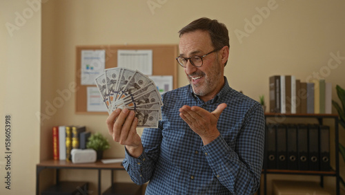 Photos Man holding cash indoors in an office setting with a joyful expression, surrounded by books and plants, showcasing financial success and happiness at work
