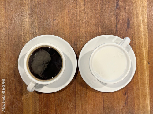 One cup of black coffee and one cup of white milk, placed side by side on a wooden table.