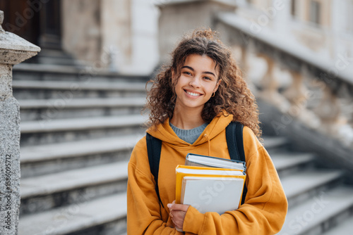 Student smiles with books on university steps during bright sunny day