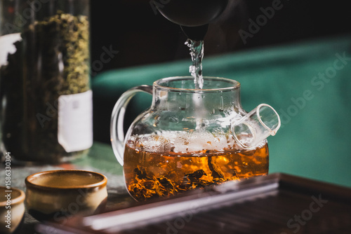 Glass teapot with loose leaf tea brewing in hot water, steam rising. Ceramic cups and tea jar on table create a cozy, rustic tea preparation scene