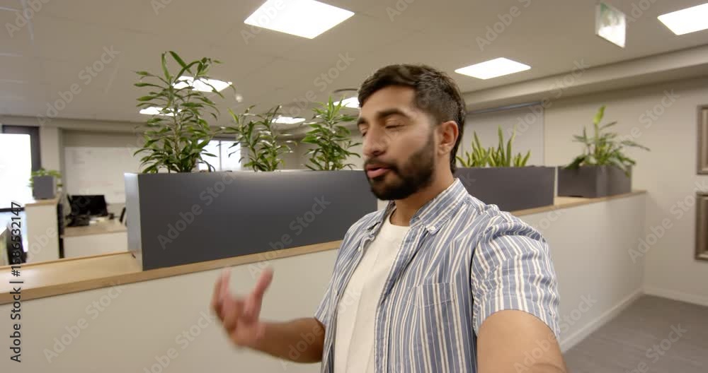 Livestreaming Indian man holding smartphone at arm's length in open-plan office, with planter boxes