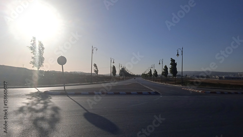 Fototapeta Naklejka Na Ścianę i Meble -  A wide-angle landscape shot of a deserted road during late afternoon or early evening, with the sun low in the sky casting a bright glare and long shadows. The road is lined with evenly spaced, tall