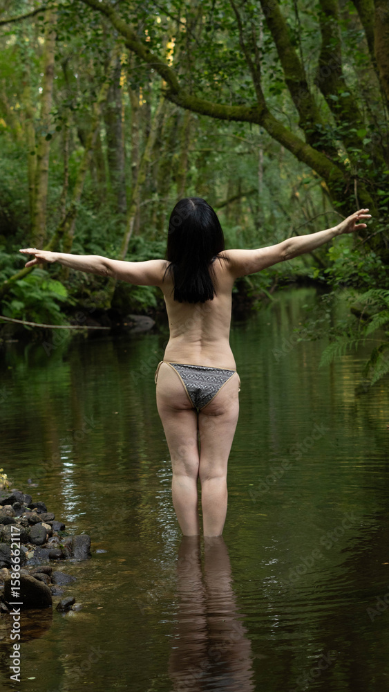 Fototapeta premium Woman on her back in a wooded stream, with arms raised in a pose of yoga or meditation. Submerged up to the thighs, it reflects peace and well-being. Dense green foliage creates a natural environment,