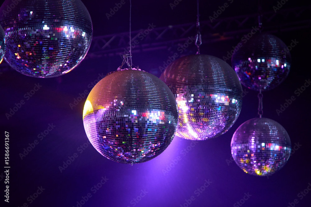 Obraz premium Fotografía artística de 5 bolas de espejos suspendidas e iluminadas con luces moradas, rosadas y doradas sobre un fondo oscuro tipo discoteca.