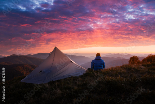 Autumn mountains glow under vibrant sunset as hiker rests near his tent. Peaceful fall scenery shown with rich evening tones and serene outdoor camping