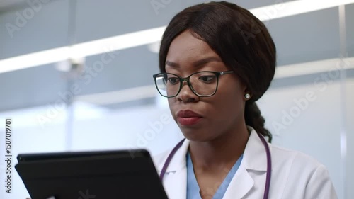 Doctor in glasses looks at a tablet, wearing a white coat and stethoscope.