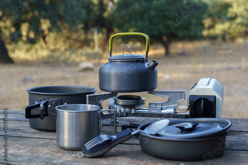 Camping kettle warming on a portable stove with various cookware items arranged on a weathered wooden table, creating a cozy outdoor cooking scene