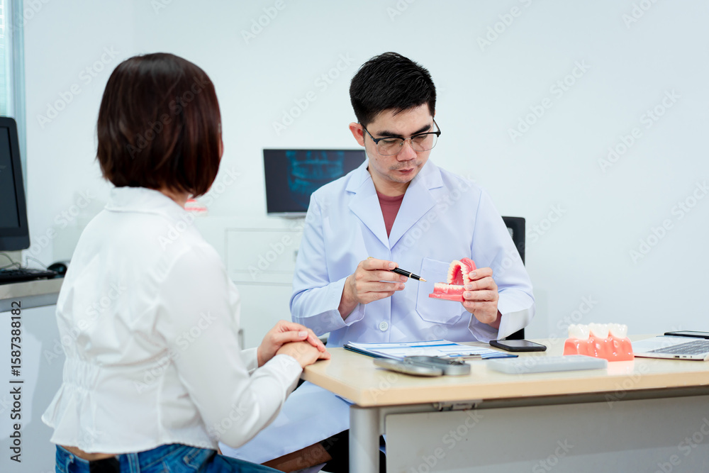 Obraz premium An Asian male dentist explains dental health to a middle-aged Asian female patient while holding a dental model, demonstrating oral care and treatment procedures in a clinical consultation setting