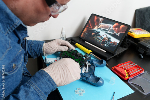 Electronic technician wearing gloves and protective glasses is repairing a video game controller using a tutorial on a tablet
