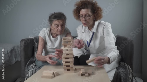 Senior woman playing Jenga, build tower of blocks. Elderly doctor in white lab coat, supporting senior patient, developing logic, fine motor skills, Alzheimer disease at home with old lonely female.