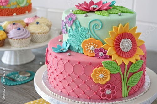 features a beautiful, two-tiered cake decorated with colorful flowers, leaves, and intricate patterns, alongside a selection of cupcakes on a cake stand.