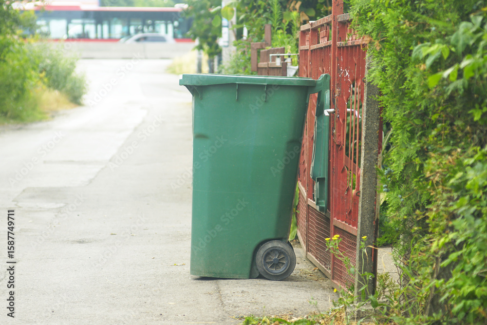 A green trash bin with an open lid stands near the fence of a house - close-up against road background. Symbol of local waste control, responsible living, environmental habits and eco-lifestyle. A green trash bin with an open lid stands near the fence of a house - close-up against road background. Symbol of local waste control, responsible living, environmental habits and eco-lifestyle.