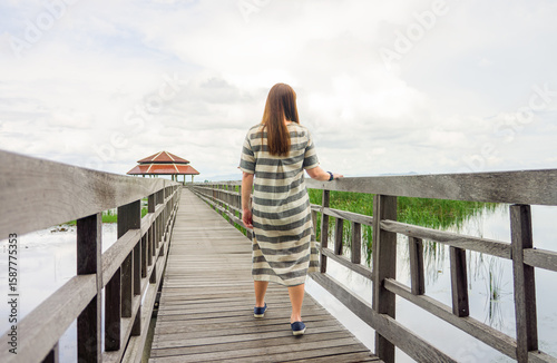 Lady Strolling on a Wooden Boardwalk Towards a Pavilion in Bueng Bua Wetland. Woman in a striped dress walks along wooden boardwalk over waters of Bueng Bua, leading towards a traditional pavilion.