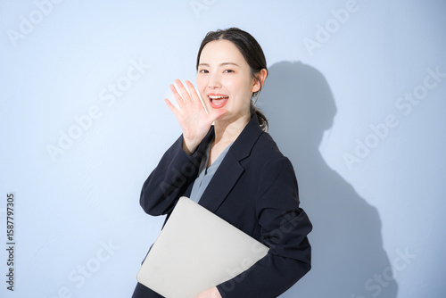 Bild auf Leinwand パソコン持ってスーツを着た立つ応援声援、内緒など呼びかける、若いかわいい女性のビジネス上半身　水色背景の営業などリクルートのイメージ