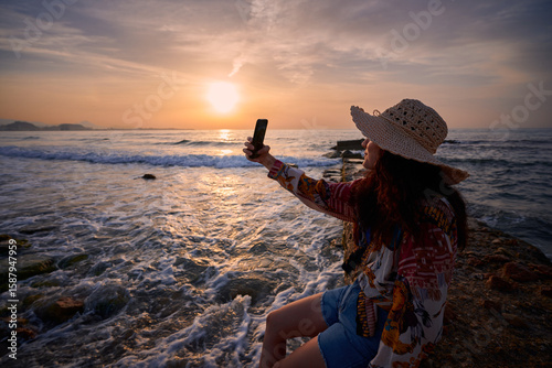 Woman enjoying sunset by the ocean while taking a selfie on the beach
