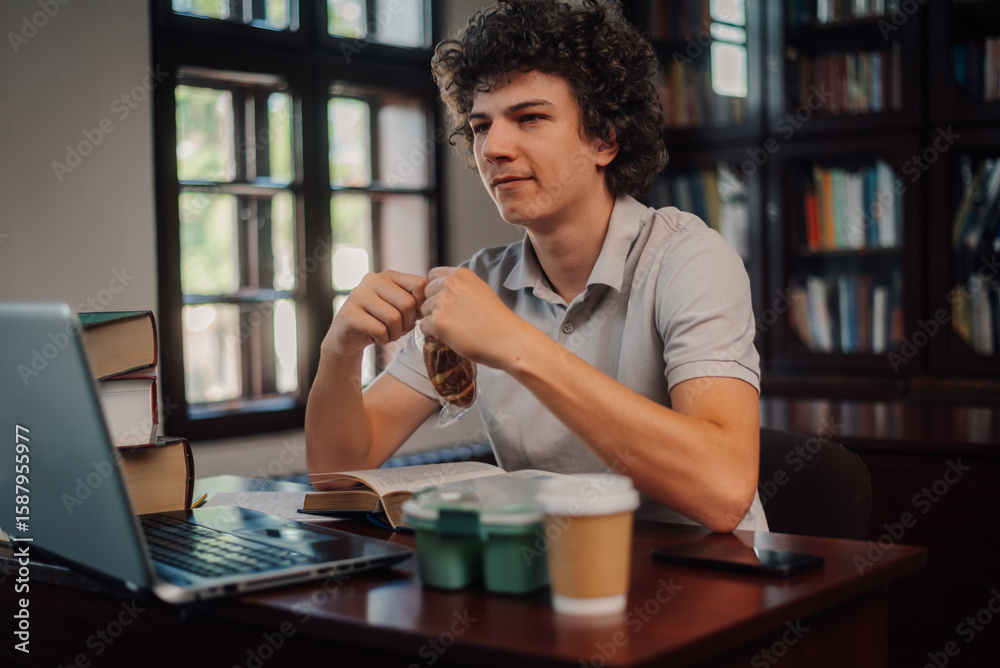 Fototapeta premium Young student eating a snack while studying in the library