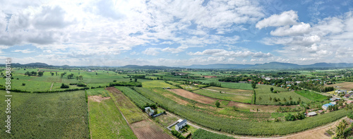 Aerial panoramic view of lush green farmland stretching across the countryside with scattered homes distant hills and a blue sky filled with soft clouds.