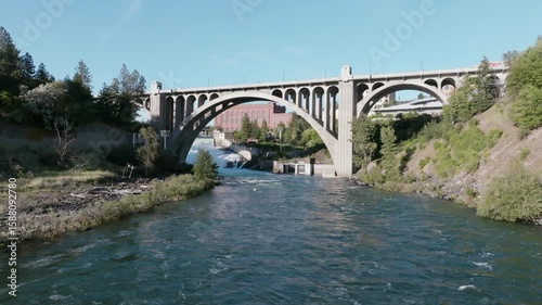 A downstream view of the Spokane River rushing beneath the Monroe Street Bridge with Spokane’s redbrick skyline and lush green banks on both sides.