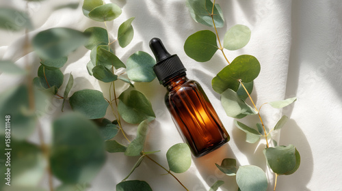 Amber bottle lies among eucalyptus on white fabric. Showcases natural essence, promotes wellness, ideal for marketing, web design, or editorial use.