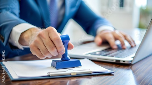 Photo of a businessman in a blue suit stamps an official document while working on a laptop in an office setting