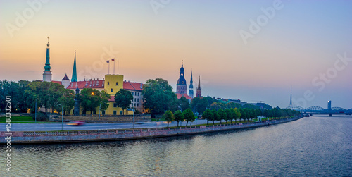 Panoramic view of Riga old town, Latvia at dusk with the Daugava River, Riga Castle, historic church spires, and city skyline under a soft pastel sky.