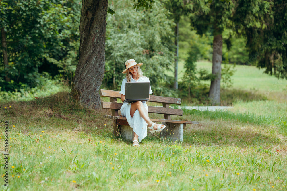 Fototapeta premium Summer vibes as woman in white dress uses laptop on bench in peaceful rural nature setting