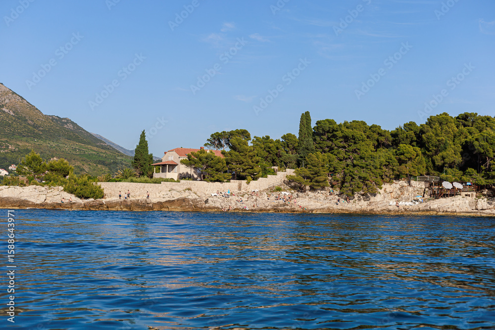 Naklejka premium Rocky coastline with people sunbathing near pine trees and a Mediterranean house.