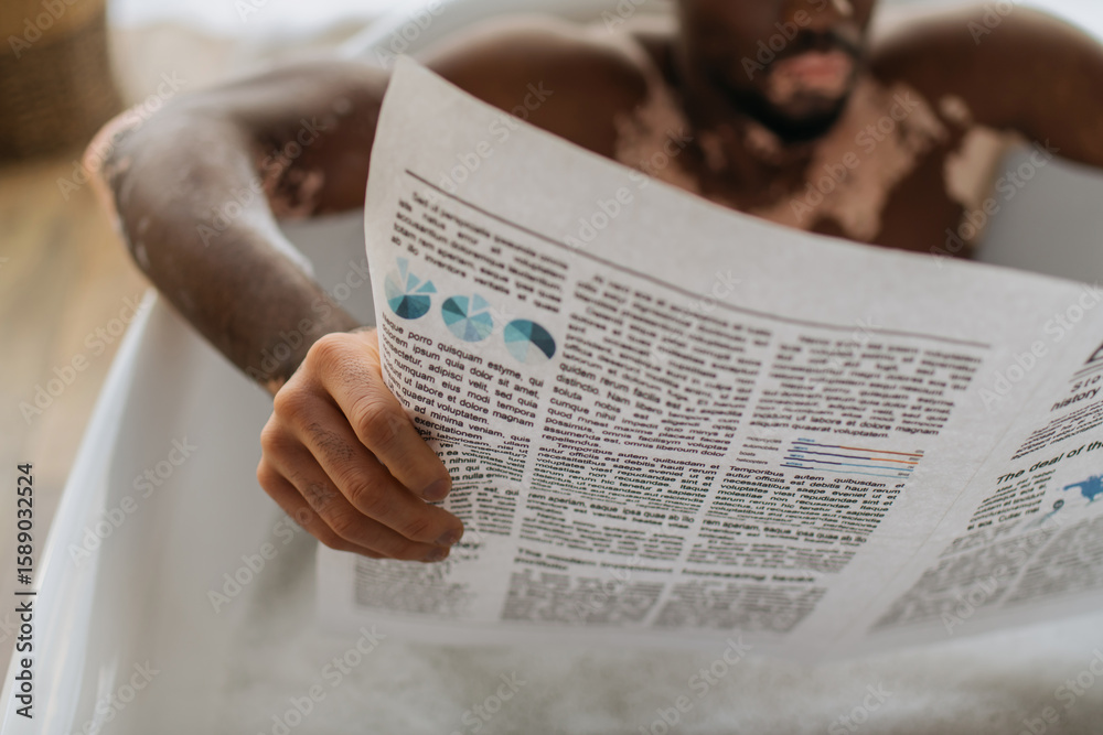 Naklejka premium Cropped view of blurred african american man with vitiligo reading newspaper while taking bath in morning