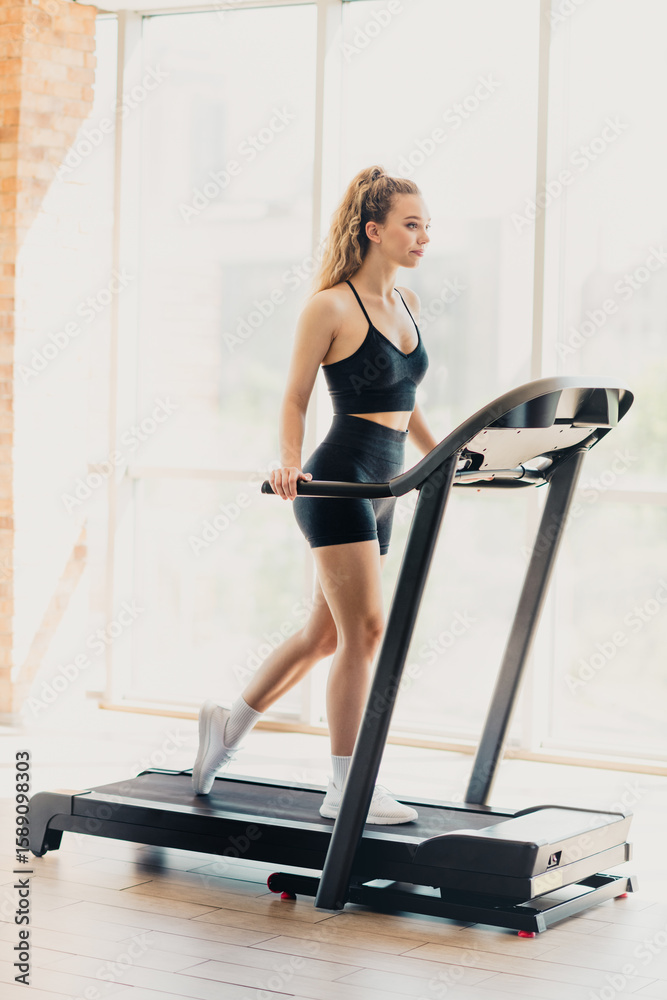 Fototapeta premium Athletic active woman exercising on a modern treadmill in a bright interior gym, practicing healthy lifestyle and sports.