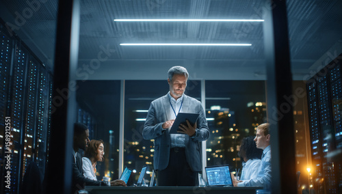 Tech team collaborates in data center at night. Leader reviews tablet. Modern, corporate look for web, marketing.