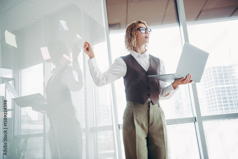 Fototapeta premium Confident businesswoman using a laptop and pointing at notes on a transparent board in a modern office