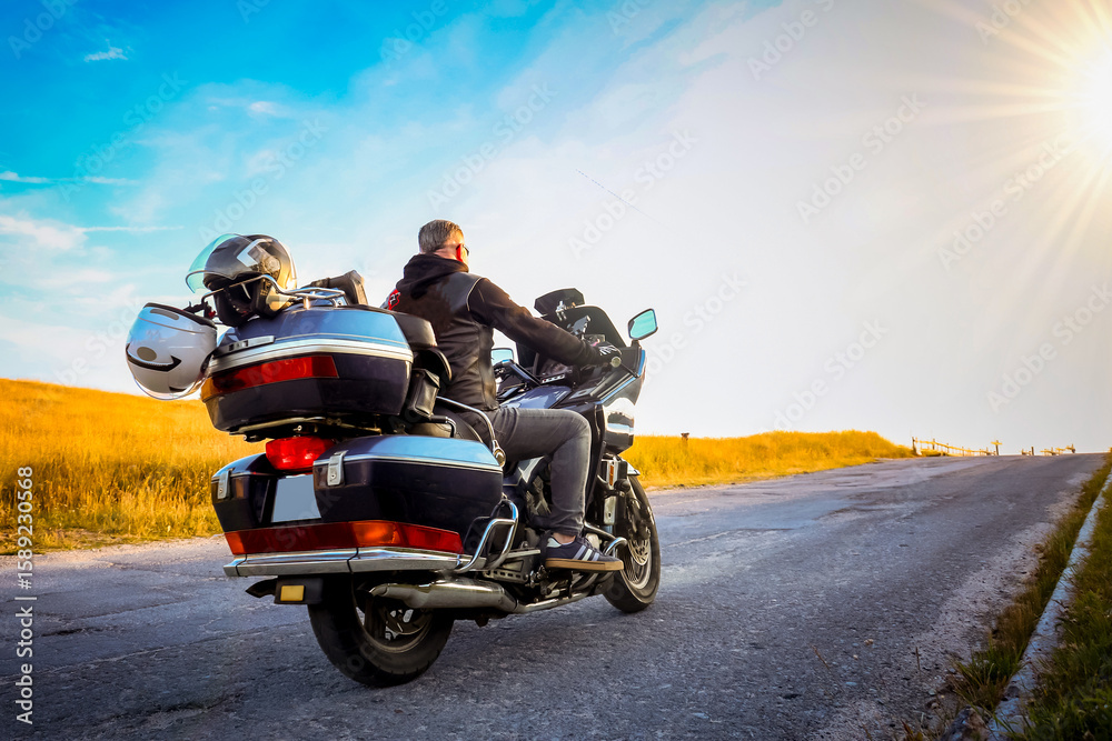 Fototapeta premium A person on a touring motorcycle riding into the sunrise on a rural road. Concept of freedom, solitude, and adventure on the open road. Space for copy on the right side.